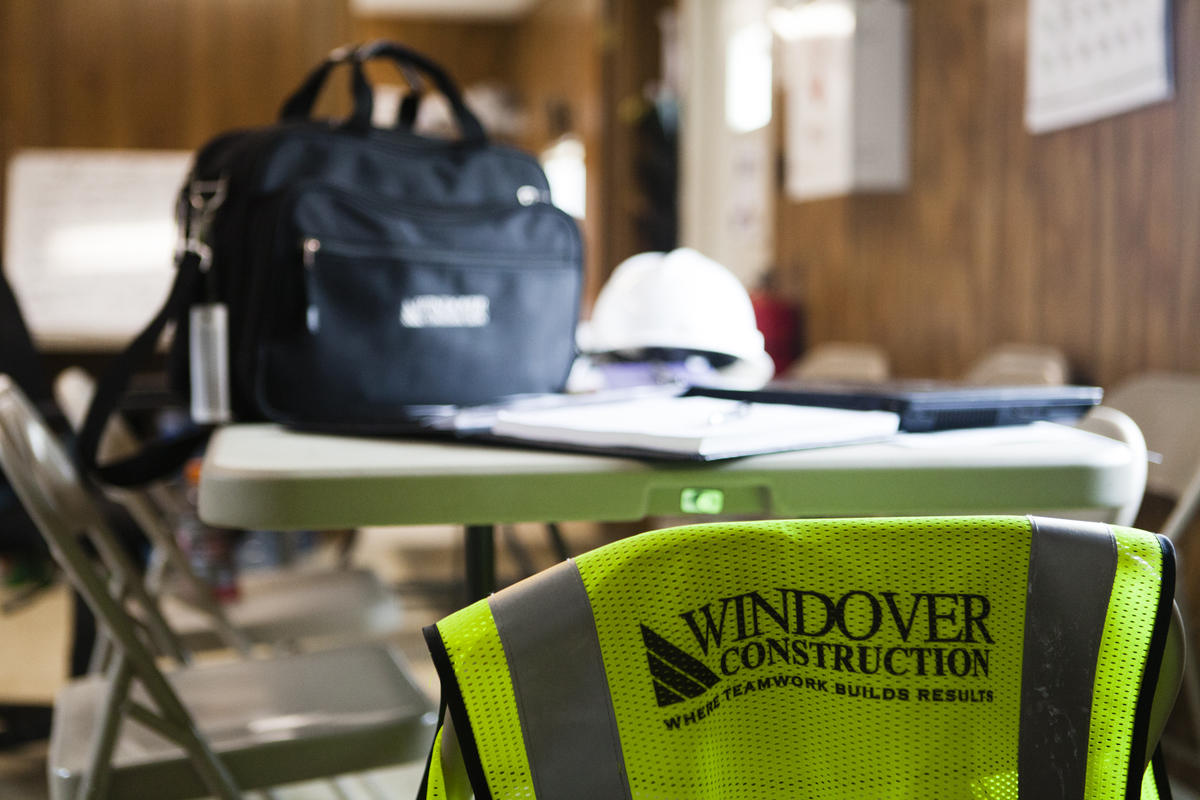 Photo of desk with Windover bag, hard hat, and vest hanging on a chair.